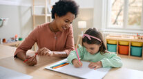 Happy black teacher and small girl drawing on paper at kindergarten.